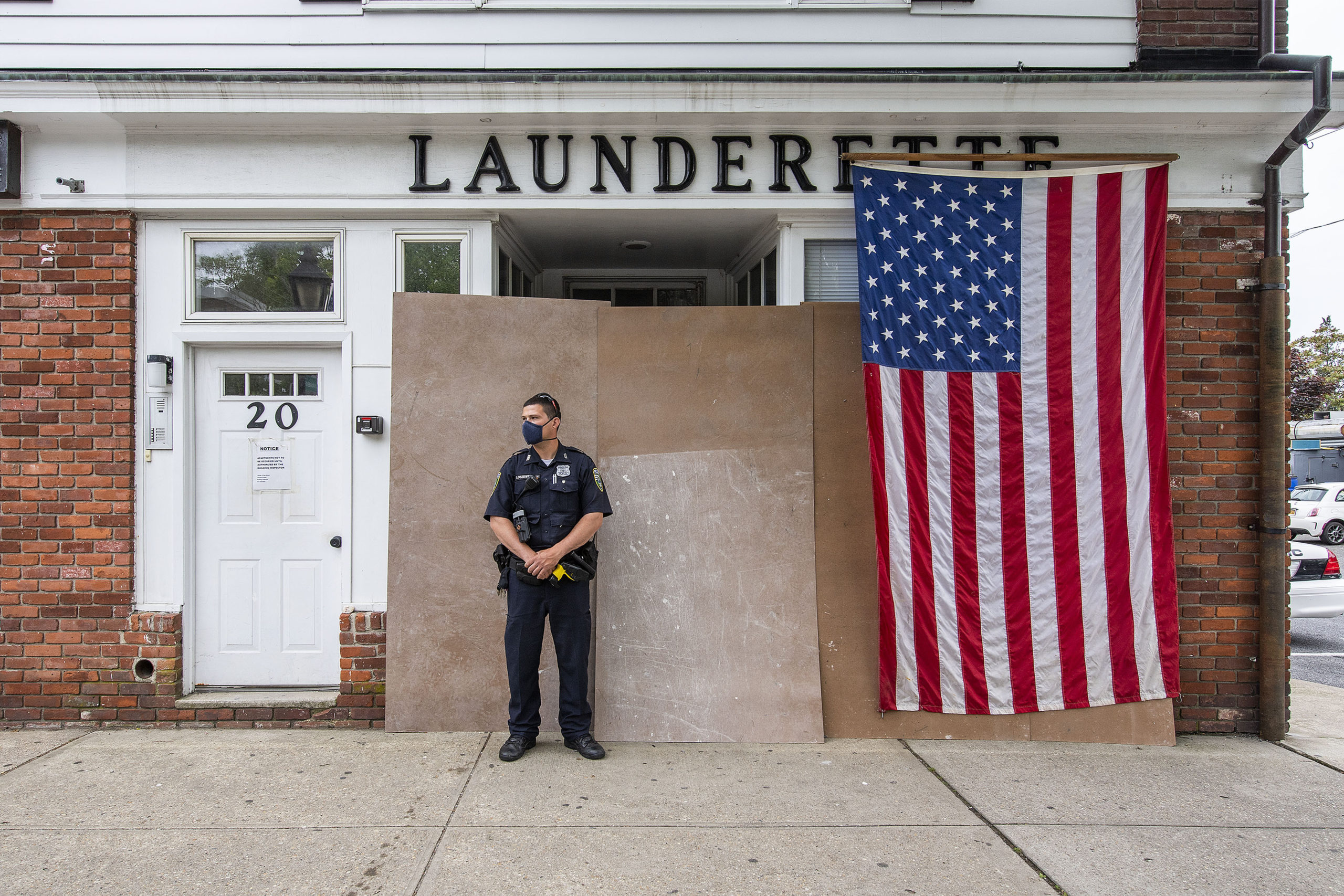 Riverhead Police Officer Patrick Loszweski watches marchers make their way down Main Street from in front of the hastily-barricaded Sag Harbor Launderette during a Black Lives Matter protest rally held in Steinbeck Park on Friday afternoon.   MICHAEL HELLER
