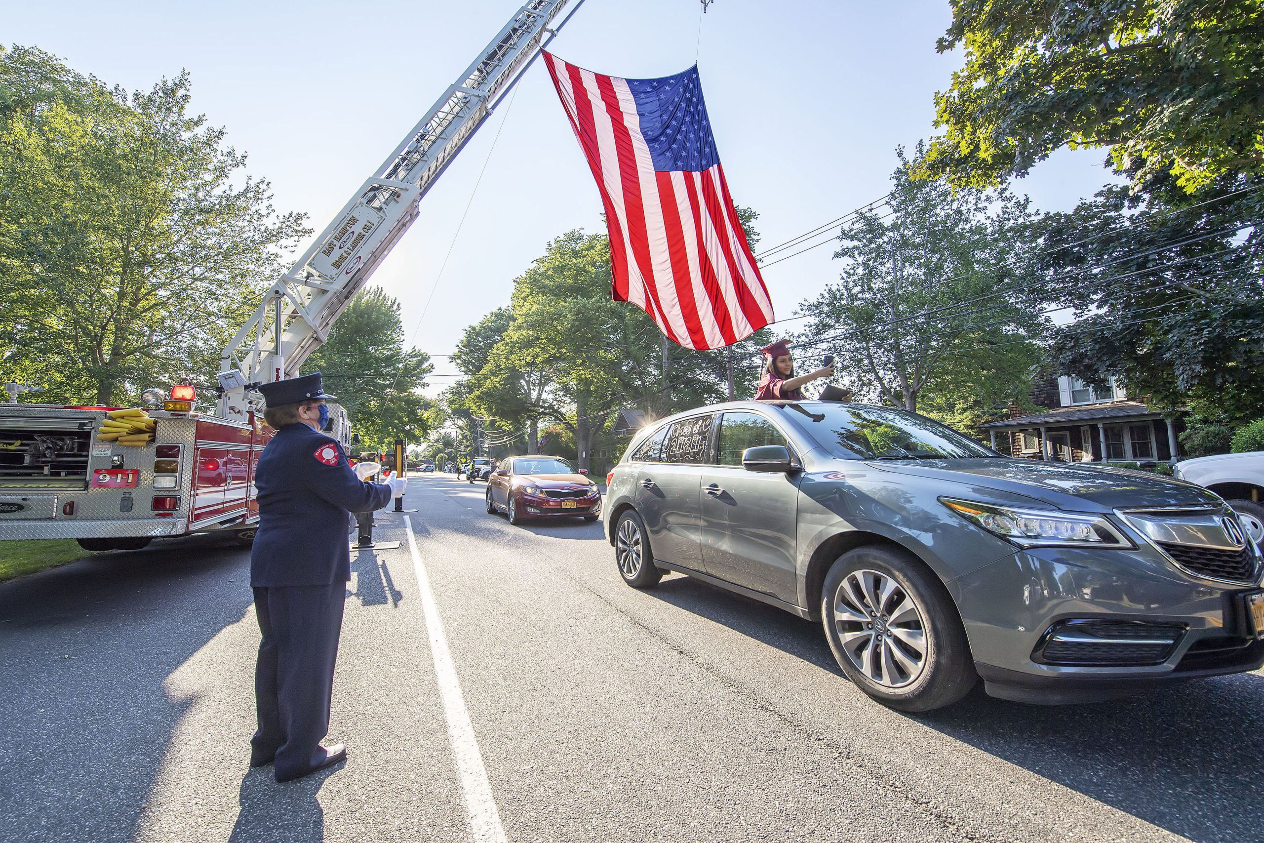A newly-graduated East Hampton High School senior gets a round of applause from East Hampton Fire Department Fire Police member Joan Jacobs as she photographs herself passing underneath a large American flag hoisted by EHFD Hool & Ladder Co.#1 in honor on the graduates on Newtown Lane following the 2020 graduation ceremony at the East Hampton High School on Friday.