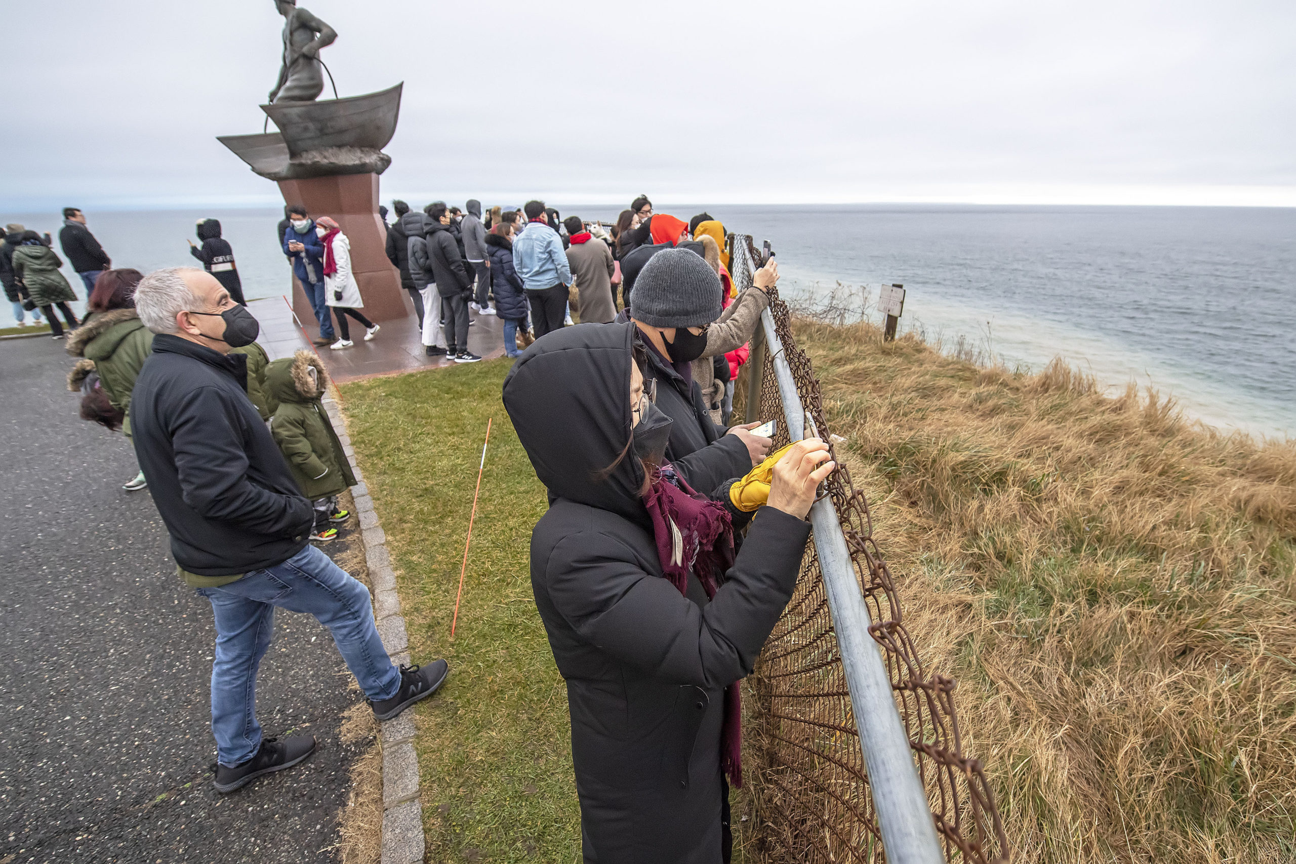 As Southampton Town Supervisor Jay Schneiderman, at left, looks on, Crara Ryu of Bayside, Queens joined a smaller-than usual crowd as she photographs the first sunrise - obscured by fog and clouds, unfortunately - of 2022 as part of a South Korean tradition, which promises good luck and wishes to those who observe it, at the Montauk Lighthouse on New Year's morning.   MICHAEL HELLER