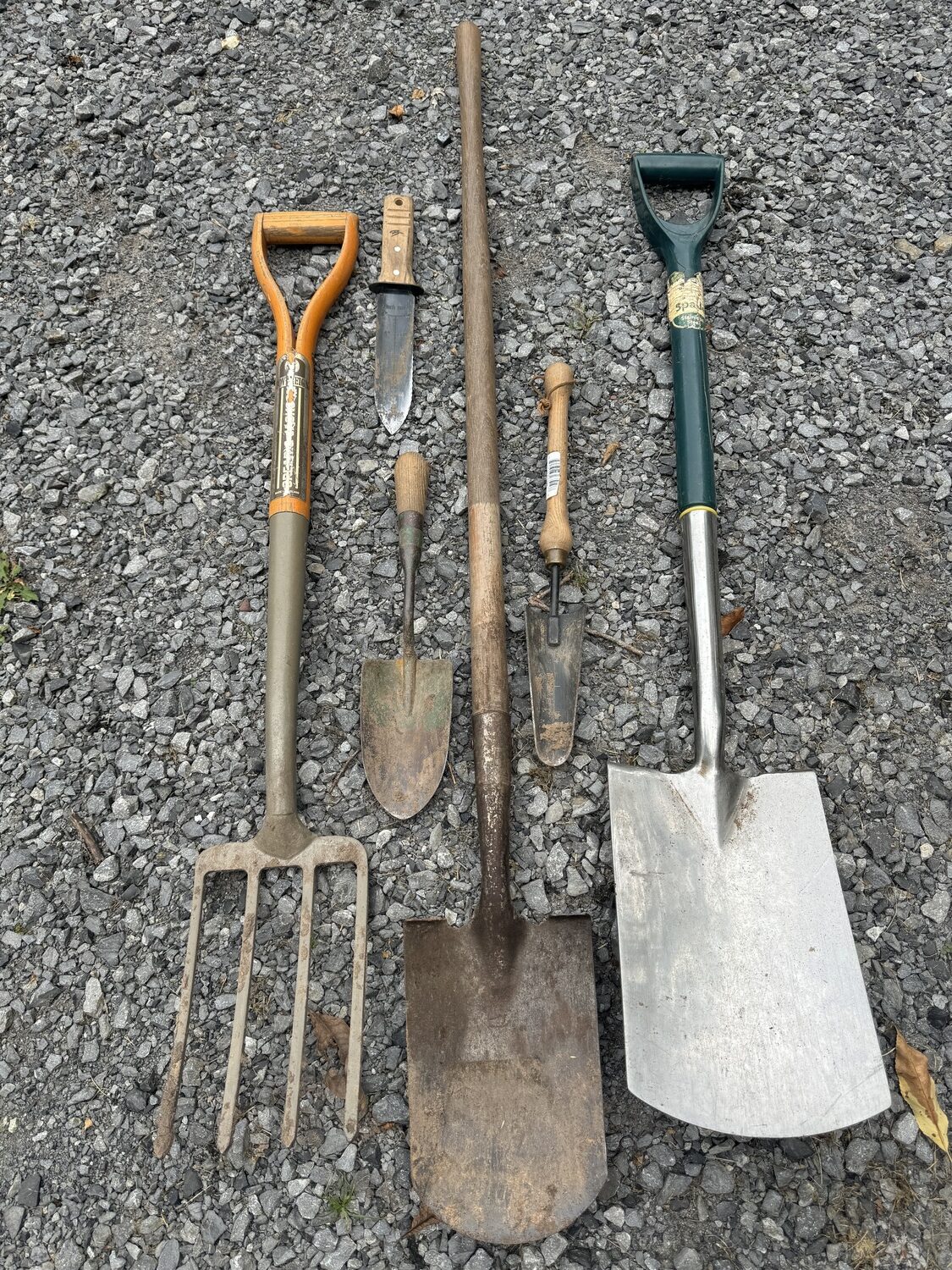 Tools you may find useful. A small digging fork on the left. Top left is a hori hori knife with a serrated edge and a cutting straight edge. Below it is a steel forged trowel. In the center with the long handle is a round-tipped straight spade. To the right is a narrow trowel, and on the far right a wide blade straight spade.  ANDREW MESSINGER