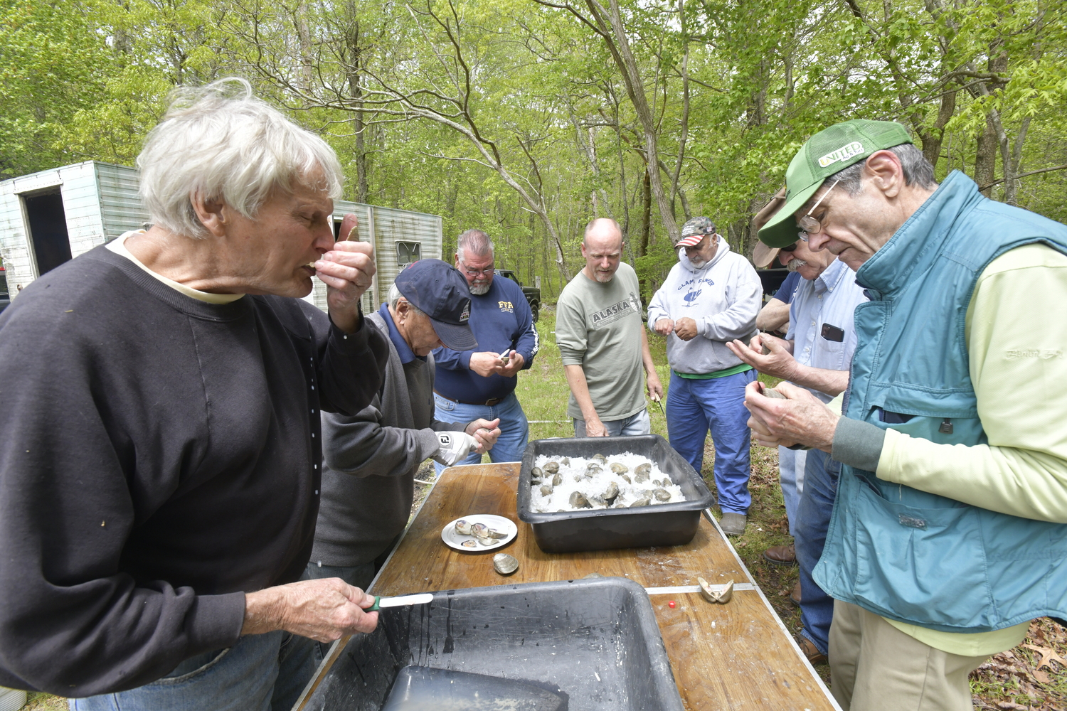 A half-dozen men surround a table on which hundreds of hardshell clams sit on ice, the men slicing them open and offering clams on the half-shell to any and all takers.  DANA SHAW