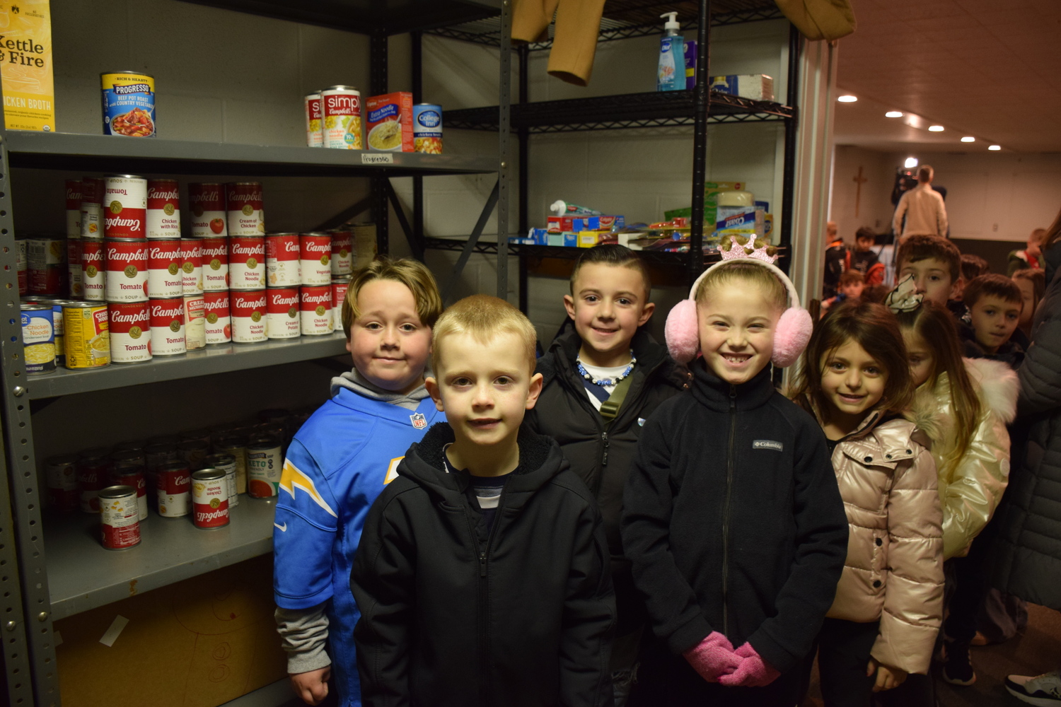 First graders at Tuttle Avenue School stocked the pantry shelves at Eastport Bible Church with 1,000 additional cans of food.  As part of a civics lesson, the students were asked to address a local issue. The first graders decided that food insecurity was a problem among the community, so they set a goal to collect 1,000 cans of food. The food drive, called the Souper Bowl, encouraged the
entire school community to participate.
Holding the collected cans, the first graders then walked to Eastport Bible Church, where they were greeted by Pastor Kaleb Kemper and administrative assistant Judy Remski, and placed their cans on the pantry’s shelves. COURTESY EASTPORT-SOUTH MANOR SCHOOL DISTRICT