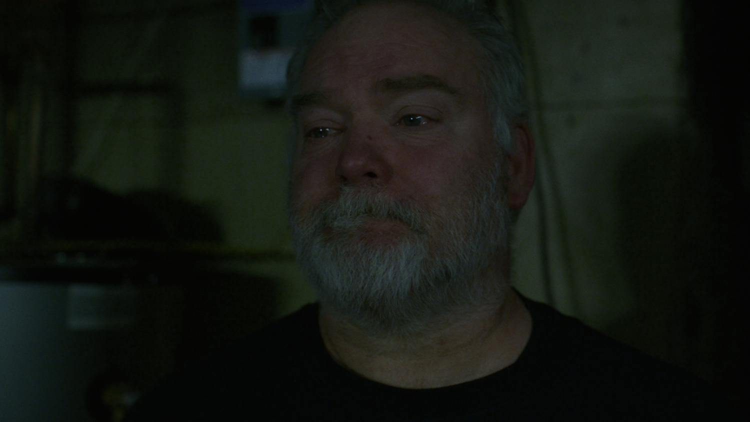 Mike Carroll stands in his dark basement with tears in his eyes and a sad expression. PRODUCTION COMPANY